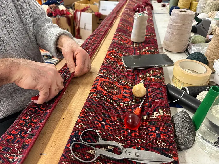 Artisan reprenant la lisière d'un tapis d'Orient sur son établi, ciseaux et bobines à portée
